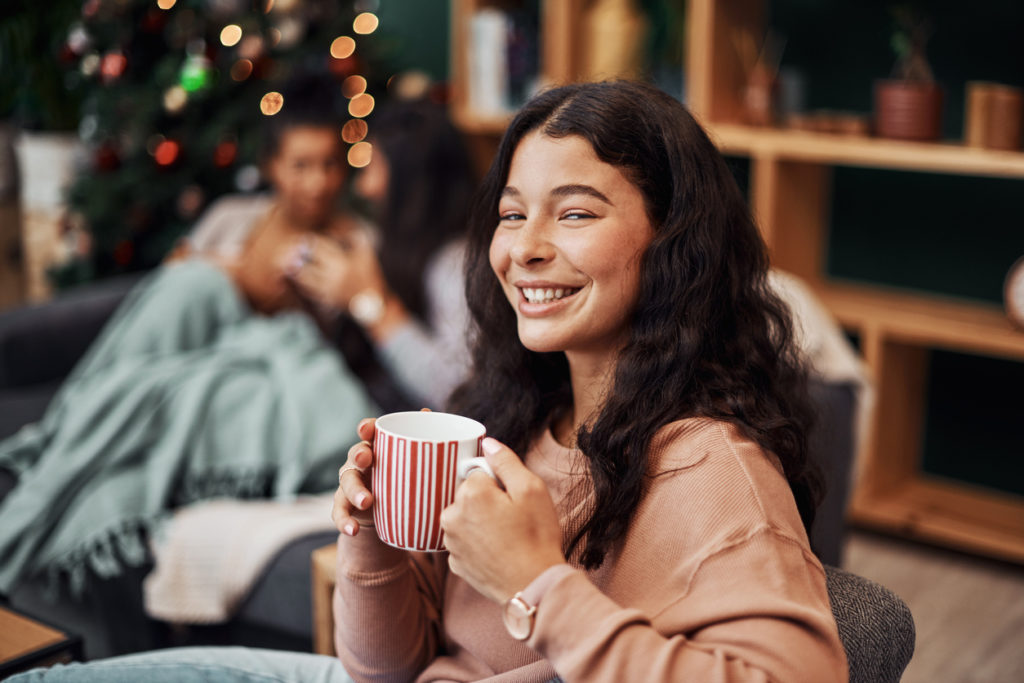 Shot of a young woman enjoying a warm beverage with her friends in the background during Christmas at home