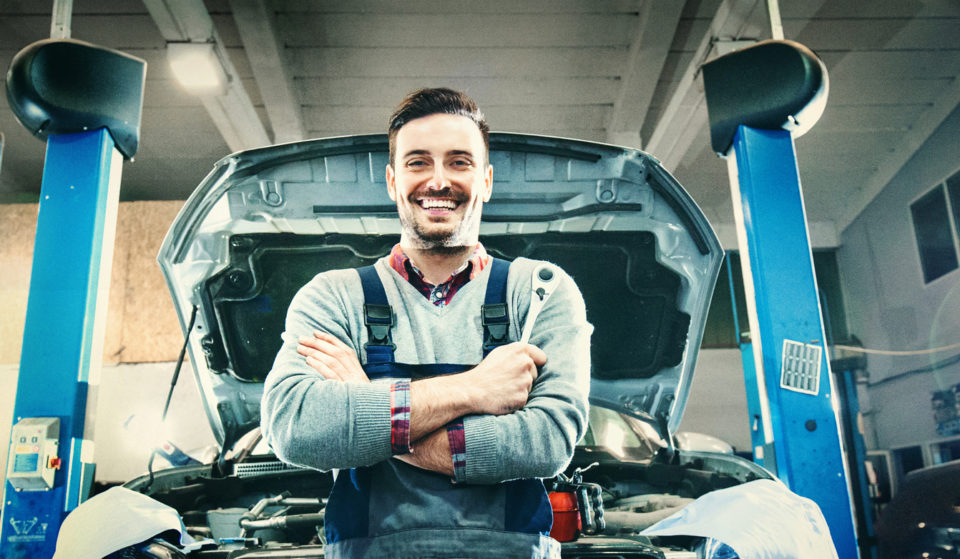 Smiling service technician standing in front of a car in his shop